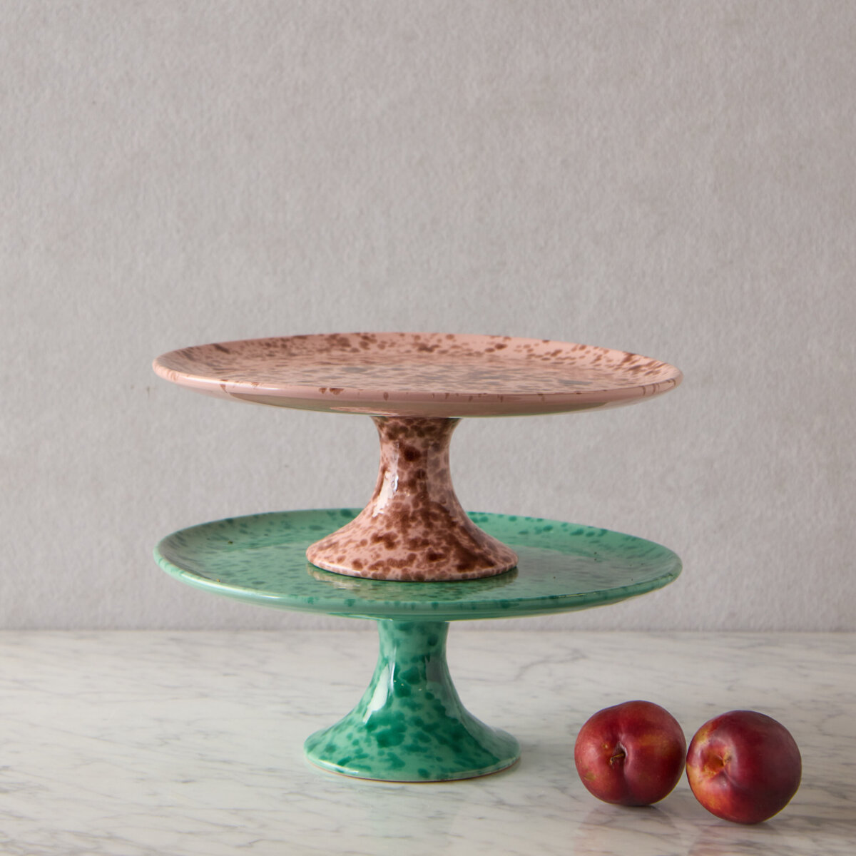 A Nashville Tastemaker on Table-Setting Do's and Don'ts, Hosting Disasters, and More - 6 Two ceramic cake stands, one pink and one green, are stacked on a marble surface next to two red plums, with a neutral backgroundβperfect for inspiring your next tablescape ideas.