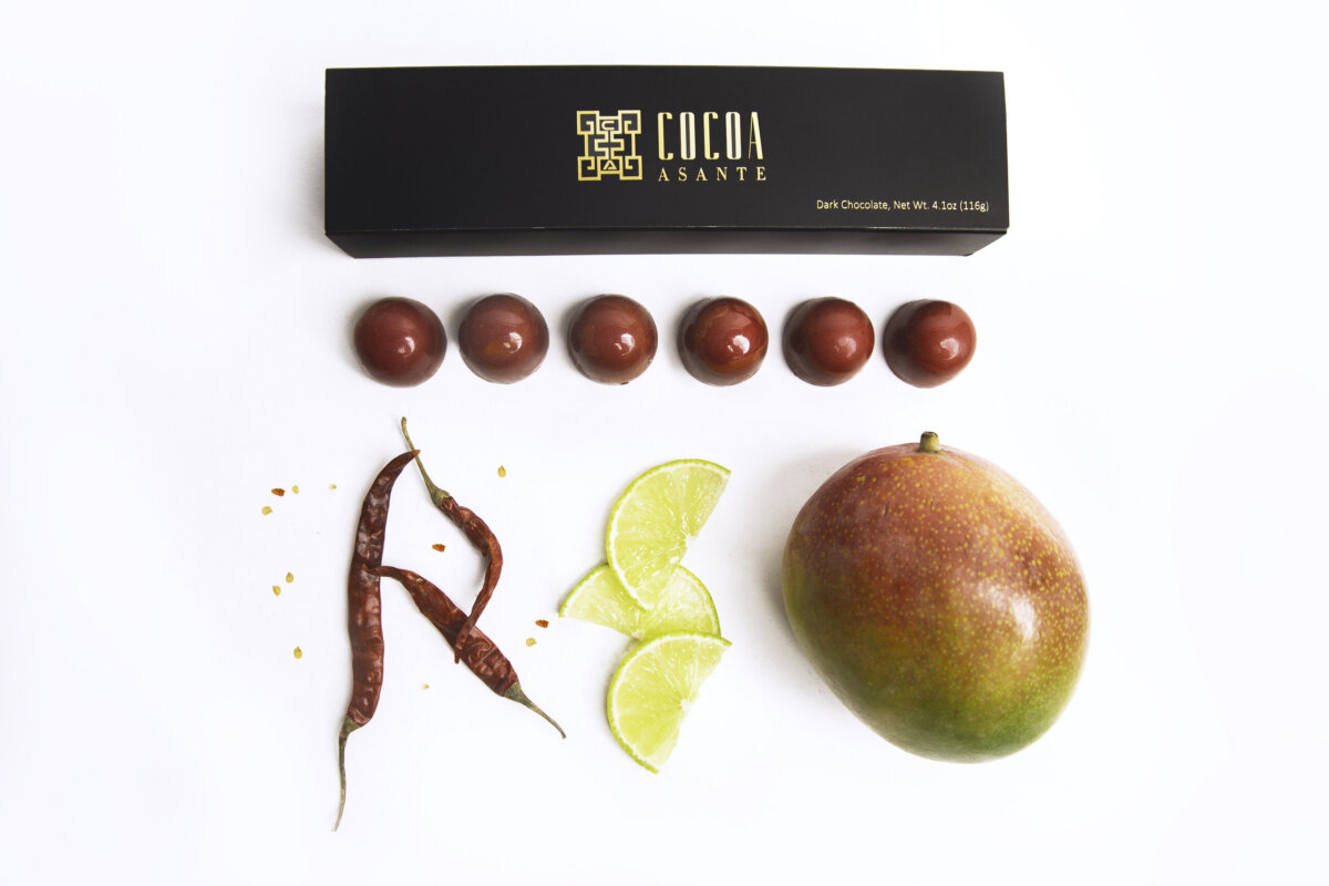 Meet the Chattanooga Chocolatier Behind Cocoa Asante - 4 A box of Cocoa Asante chocolates, six red chocolate spheres, two dried red chilies, three lime slices, and a mango arranged on a white background.