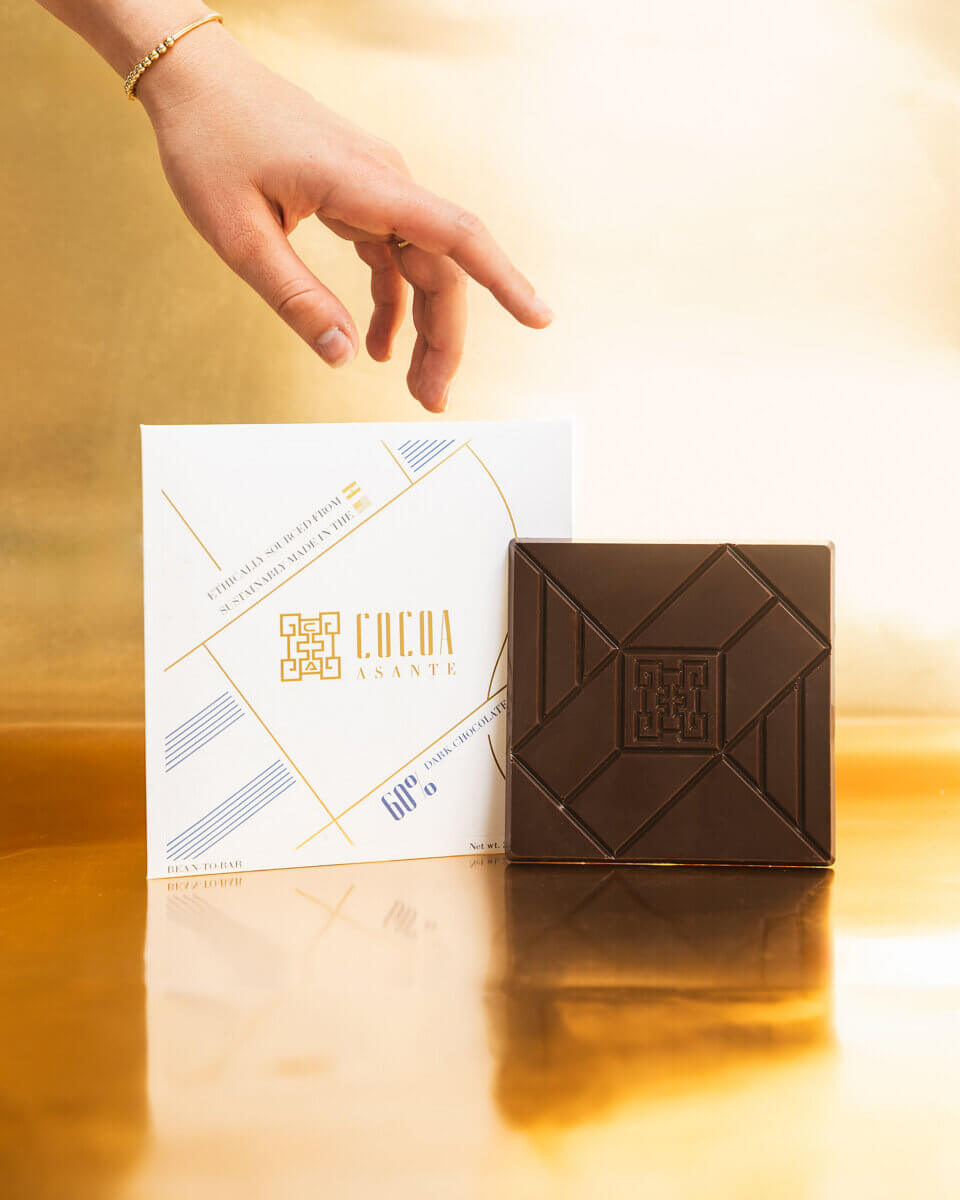 Meet the Chattanooga Chocolatier Behind Cocoa Asante - 5 A hand reaches toward a square chocolate bar placed in front of a white Cocoa Asante box, designed by Ella Livingston, on a reflective gold surface.