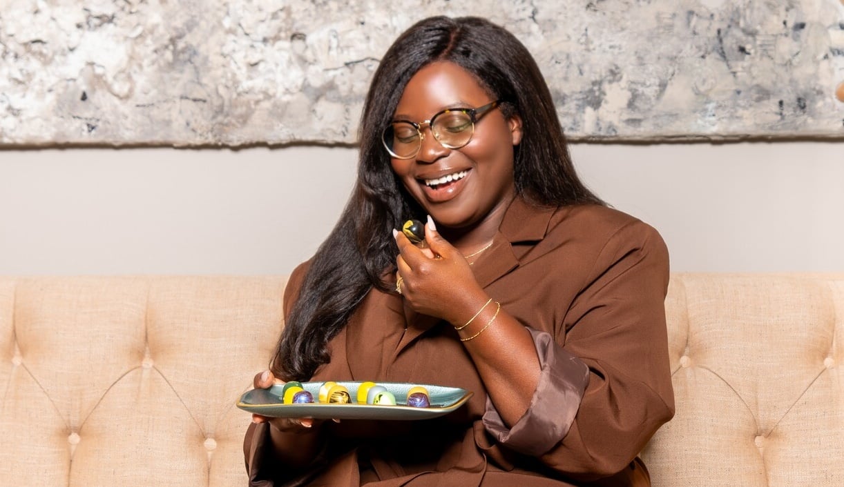 A woman in a brown outfit sits on a beige couch, smiling and holding a plate of colorful chocolates, about to eat one.