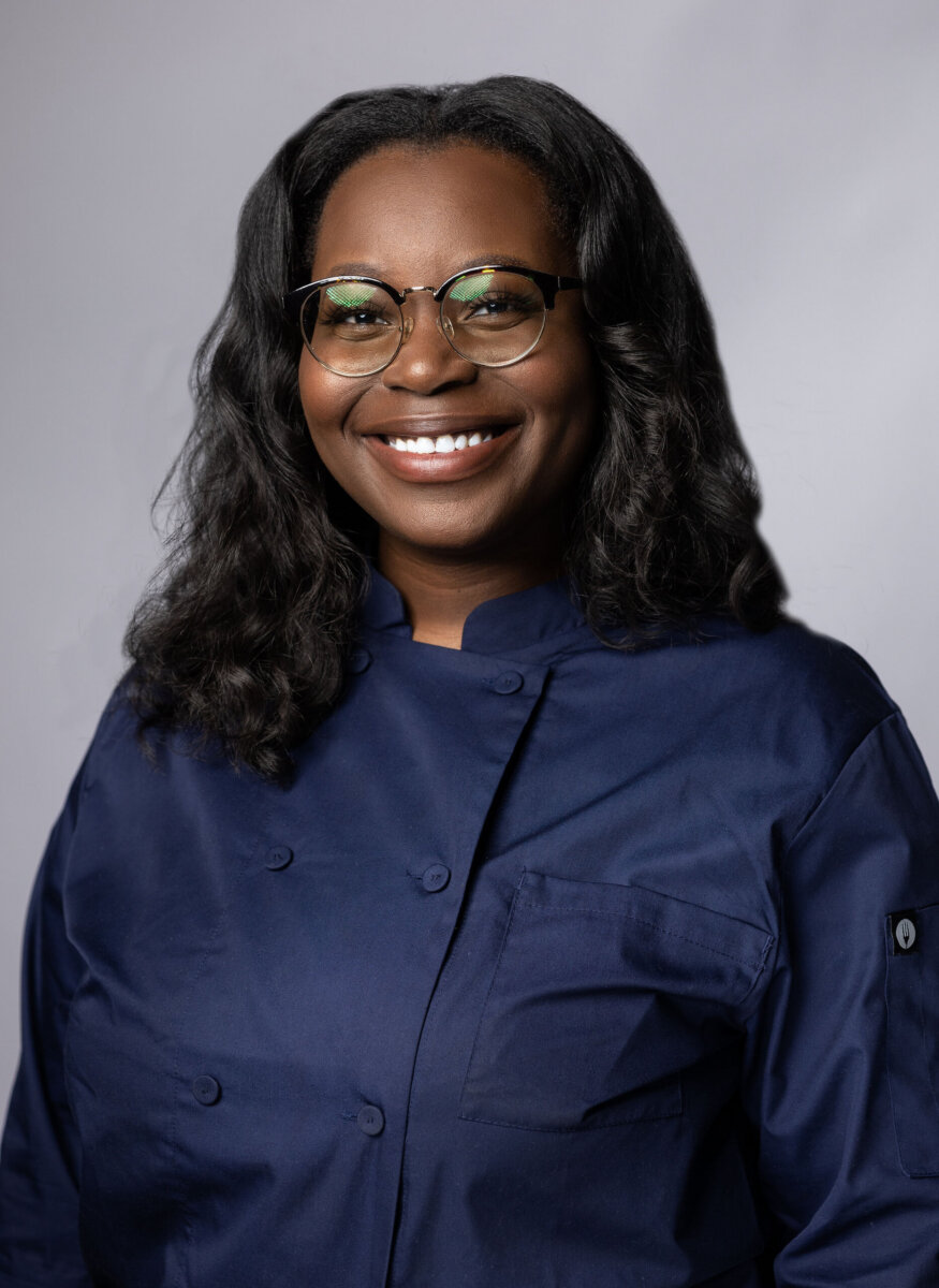 Meet the Chattanooga Chocolatier Behind Cocoa Asante - 2 A woman wearing glasses and a navy chef's coat smiles at the camera against a light gray background.
