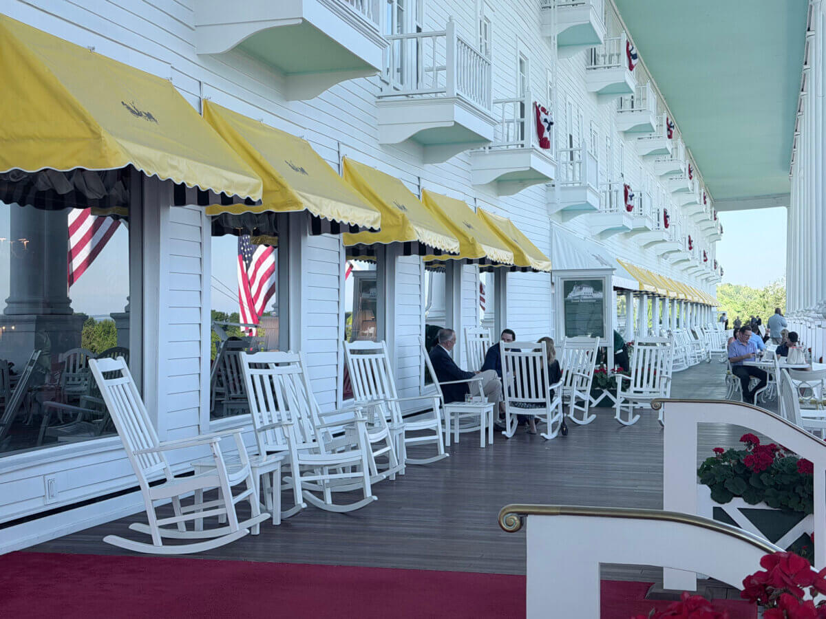Midwest Weekend Getaway: My Mackinac Island Escape - 14 White porch with yellow awnings, white rocking chairs, people sitting, large windows, and American flags reflecting in the glass. Red flowers are visible in the foreground.