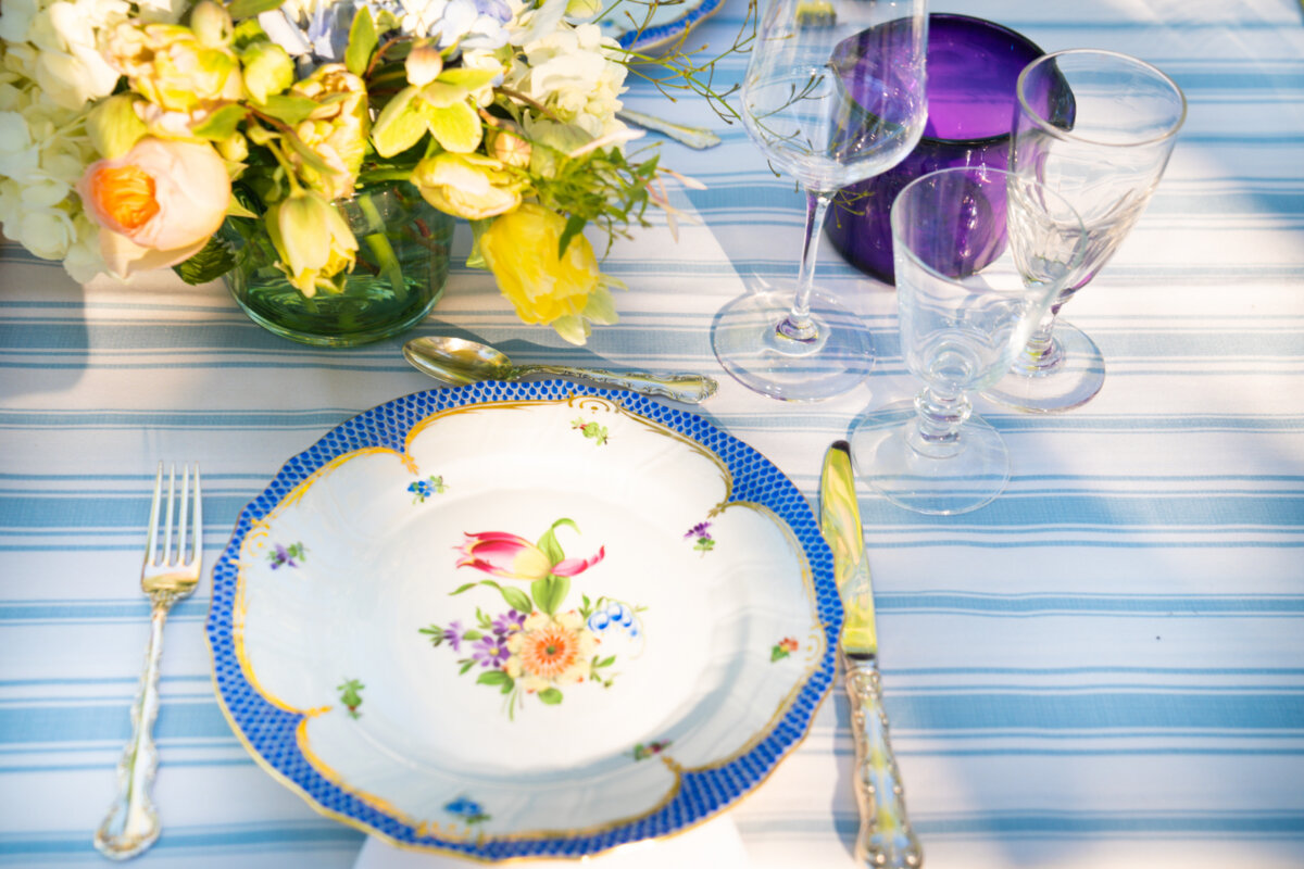 A Nashville Tastemaker on Table-Setting Do's and Don'ts, Hosting Disasters, and More - 3 A floral-patterned plate, cutlery, glassware, and a flower arrangement are set on a blue and white striped tablecloth in sunlight, offering fresh inspiration for elegant tablescape ideas.