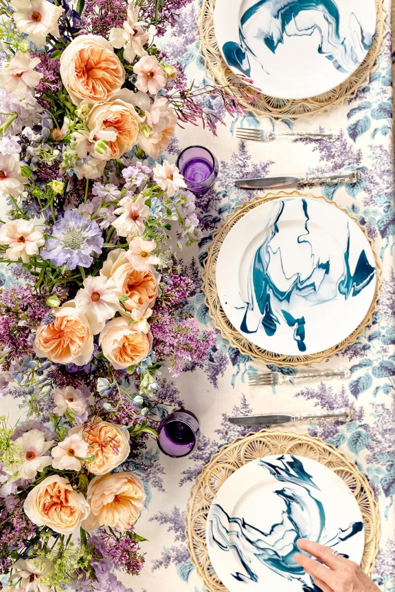 A Nashville Tastemaker on Table-Setting Do's and Don'ts, Hosting Disasters, and More - 4 Floral centerpiece with peach and purple flowers on a patterned tablecloth, surrounded by plates with blue abstract designs, woven placemats, and purple glasses. A hand reaches for a plate in this inspiring displayβperfect for anyone seeking fresh tablescape ideas.