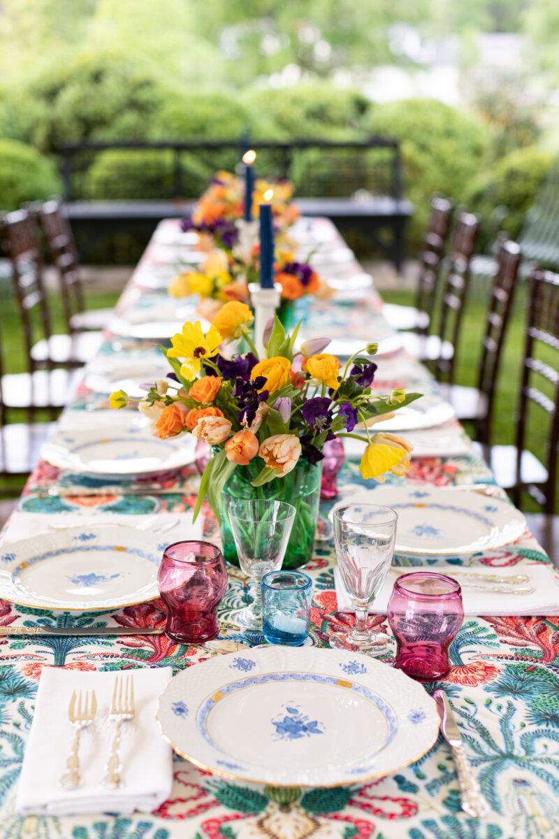 A Nashville Tastemaker on Table-Setting Do's and Don'ts, Hosting Disasters, and More - 7 A long outdoor table set with a floral-patterned tablecloth, colorful flowers, blue candles, glassware, and plates, surrounded by wooden chairsβperfect inspiration for elegant tablescape ideas.
