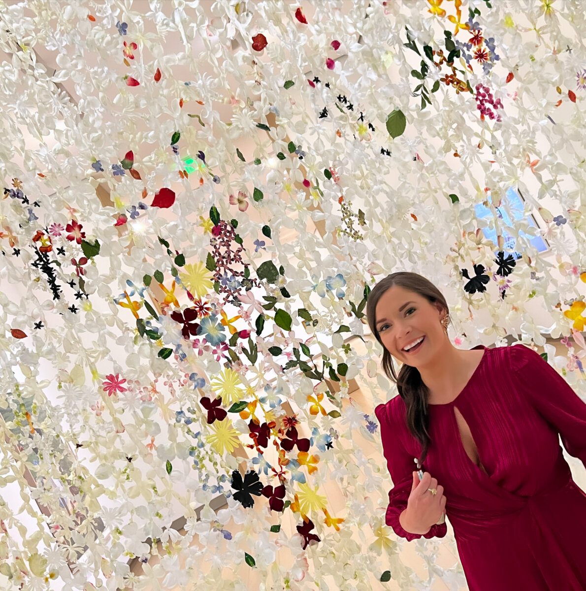 3 Southern Women Changing the Way We Travel - 7 A Southern woman in a maroon dress stands smiling in front of an art installation featuring hanging white and colorful flowers, embracing the beauty of travel and changing the way we explore new places.