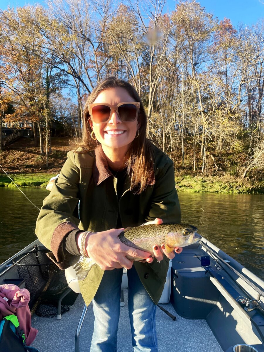 3 Southern Women Changing the Way We Travel - 6 A Southern woman in sunglasses and a green jacket holds a fish while standing on a boat, surrounded by trees and waterβchanging the way we travel, one adventure at a time.
