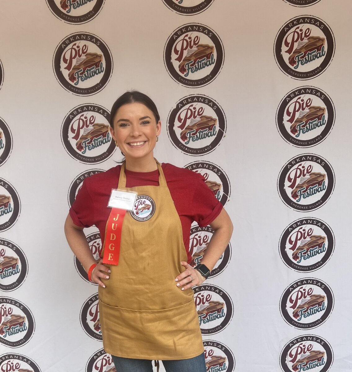 3 Southern Women Changing the Way We Travel - 5 A Southern woman wearing a gold apron and red judge ribbon stands in front of a backdrop with multiple Arkansas Pie Festival logos, celebrating local flavor and changing the way we travel through unique culinary experiences.