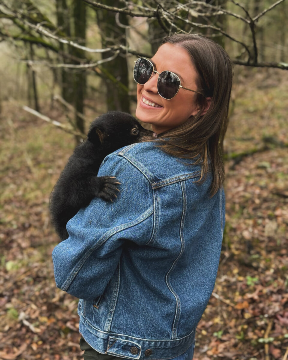 3 Southern Women Changing the Way We Travel - 4 A Southern woman in a denim jacket holds a small black bear cub on her shoulder in a forested area, embracing travel and changing landscapes.