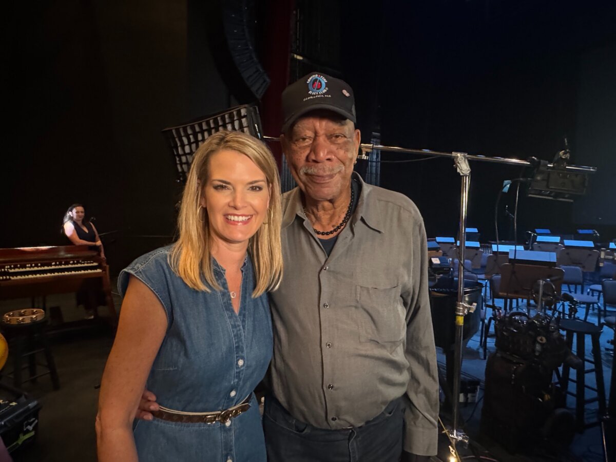 3 Southern Women Changing the Way We Travel - 2 A Southern woman in a blue dress stands next to an older man in a black shirt and cap, smiling on stage with musical instruments and equipment in the background, capturing the spirit of travel and adventure.