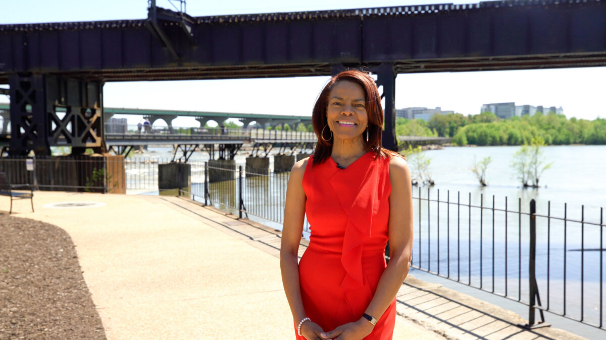 A Southern woman in a sleeveless red dress stands outdoors near a river, with a black metal bridge and city buildings in the background, capturing the spirit of travel and adventure.