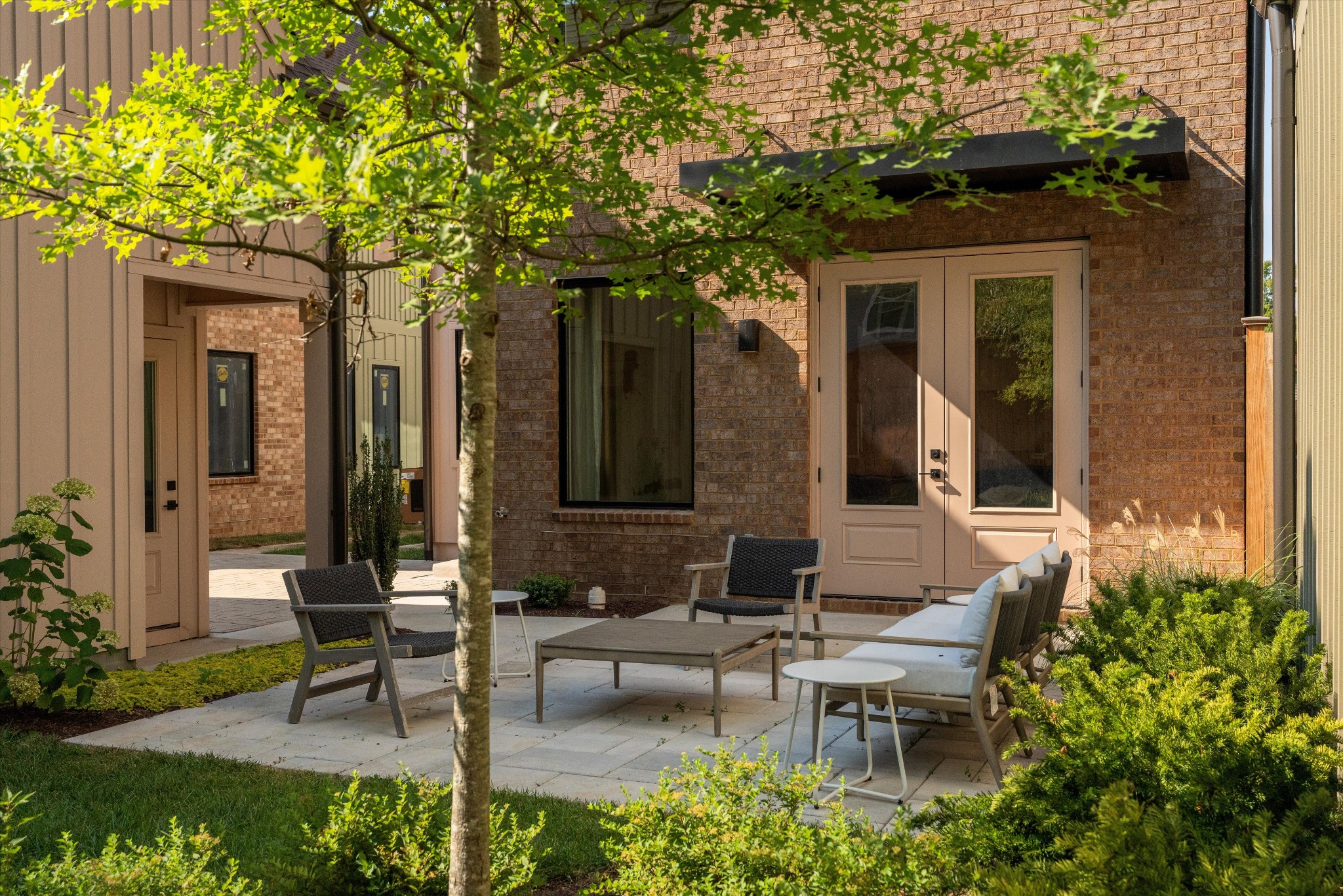 627 Richmond Bend, Nashville TN - 1 A small patio with outdoor seating and a table is surrounded by greenery and a young tree, set against the brick exterior of a house at 627 Richmond Bend in Nashville.