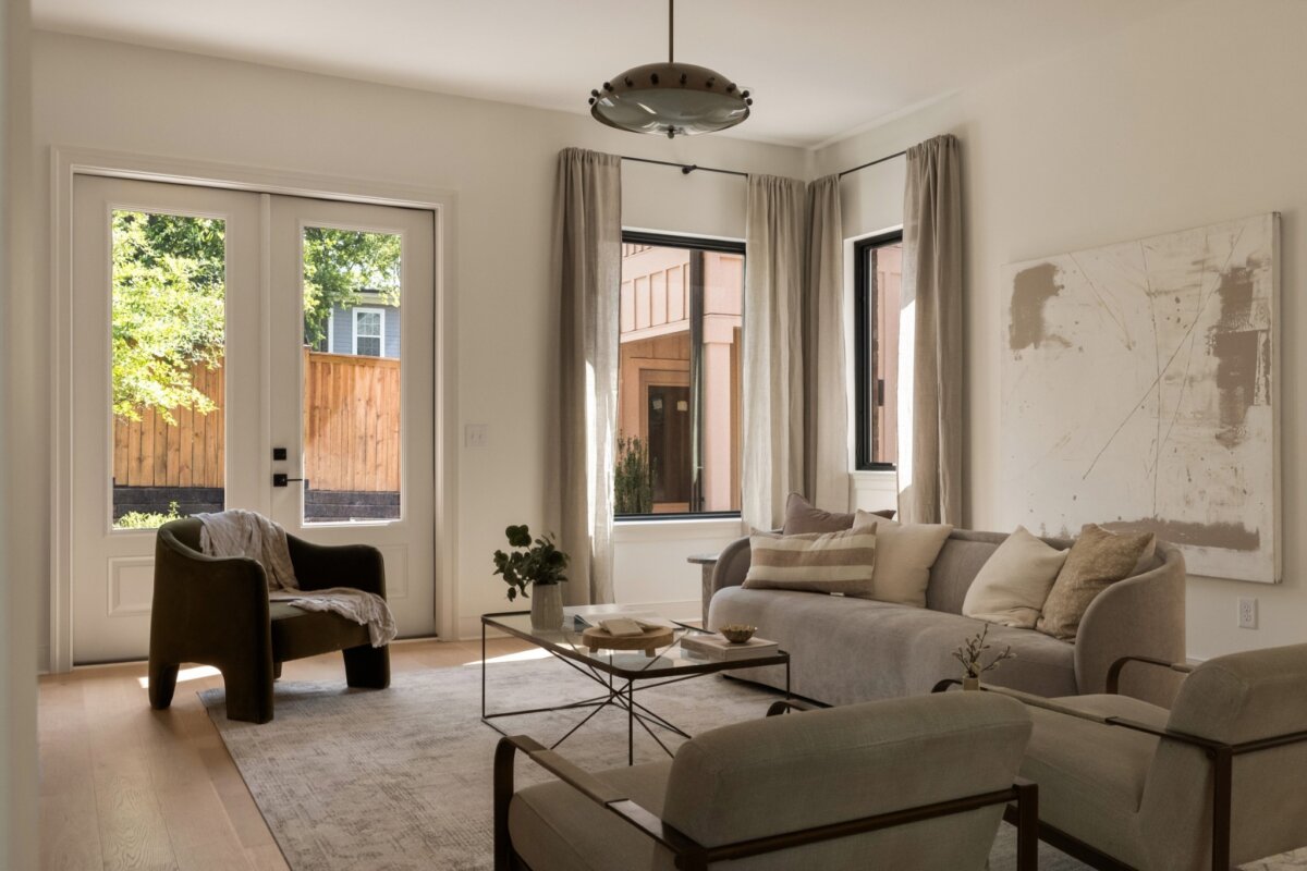 Neutral-toned living room at 627 Richmond Bend, Nashville TN, with a sofa, two chairs, a coffee table, wall art, and large windows with curtains letting in natural light. A door opens to a fenced backyard.