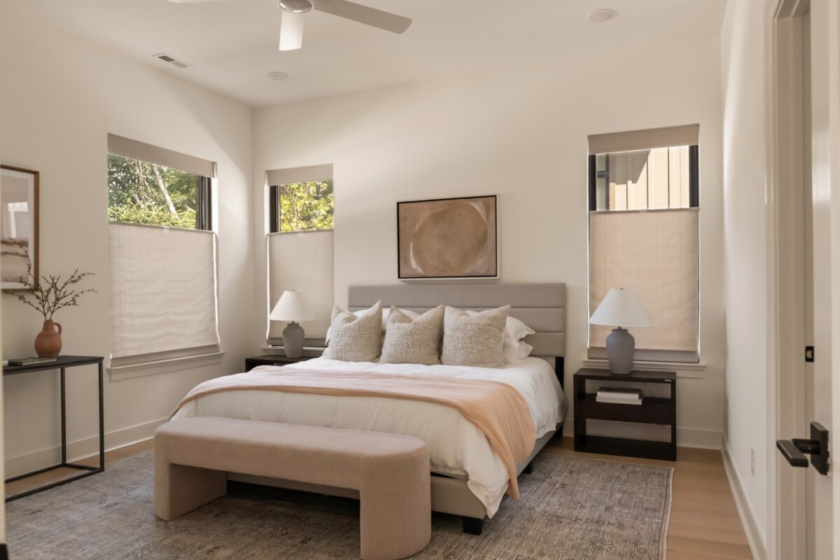 Modern bedroom in 627 Richmond Bend, Nashville, with a bed, three windows with beige shades, two nightstands with lamps, a bench at the foot of the bed, and neutral decor.