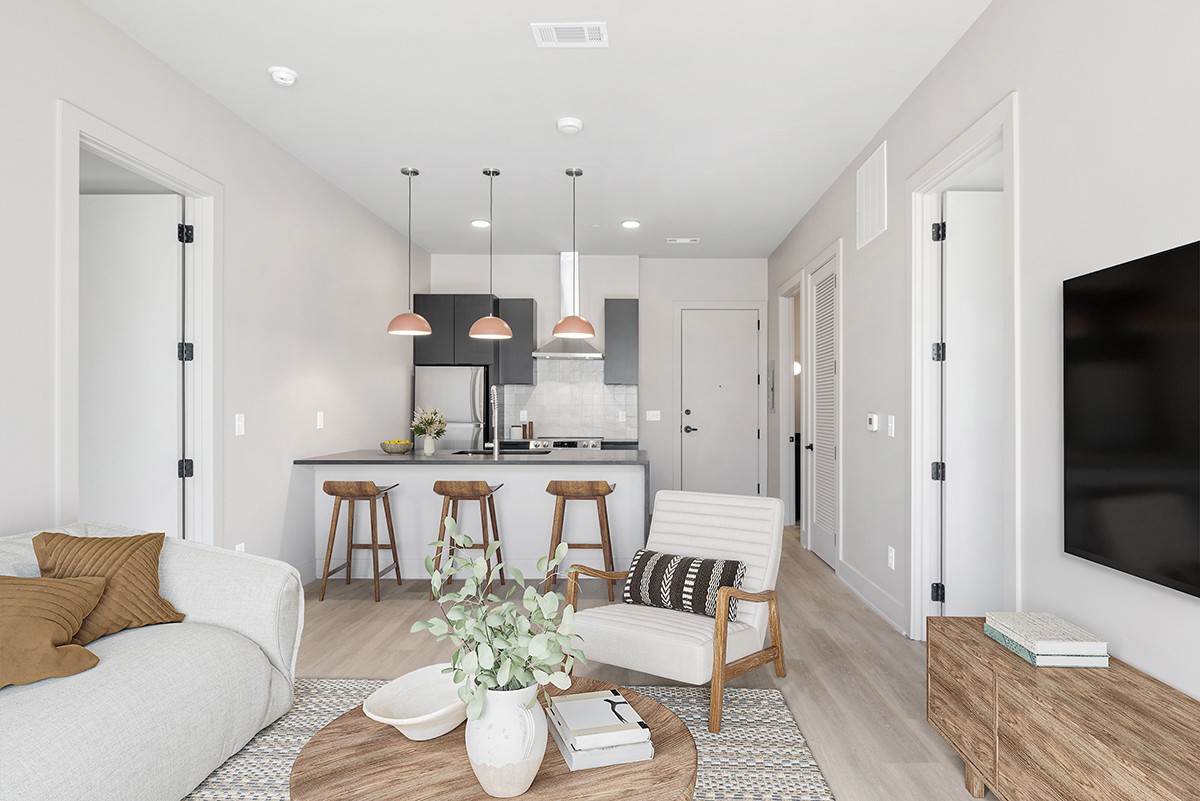Modern open-plan living area in Nashville’s desirable 1090 Haysboro Ave, featuring a stylish kitchen with barstools, a cozy armchair, sofa, coffee table with decor, pendant lights, and a wall-mounted TV. Light-neutral colors and minimalist style create an inviting atmosphere.