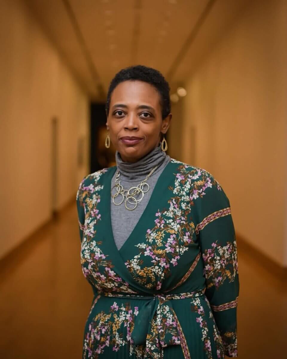 Meet Birmingham’s First Poet Laureate, Salaam Green - 2 Salaam Green stands in a hallway wearing a floral dress, gray turtleneck, and gold necklace, looking at the camera with a neutral expression.