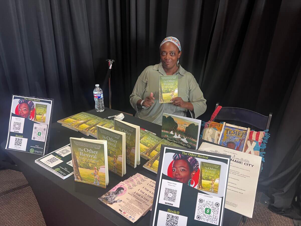 Meet Birmingham’s First Poet Laureate, Salaam Green - 3 Salaam Green sits at a table displaying books, flyers, and QR codes at an indoor event. The backdrop is a black curtain. The person, identified as Salaam Green—Birmingham’s Poet Laureate—is holding up one of the books and facing the camera.
