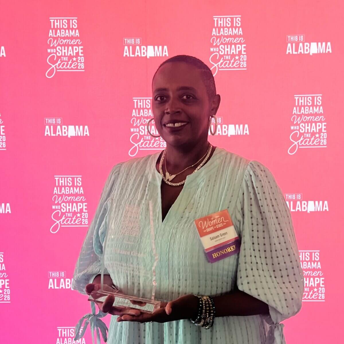 Meet Birmingham’s First Poet Laureate, Salaam Green - 7 A woman in a light blue dress holds an award in front of a pink backdrop that reads “This is Alabama Women Who Shape the State.” Salaam Green, the Birmingham Poet Laureate, wears an honoree badge and smiles at the camera.