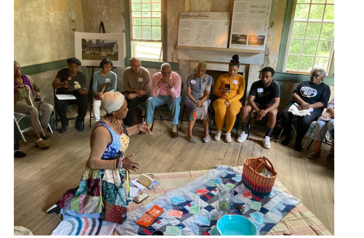 Meet Birmingham’s First Poet Laureate, Salaam Green - 5 A woman sits on a blanket surrounded by cards and objects, speaking to a diverse group of adults seated in a circle in a well-lit room with tall windows.