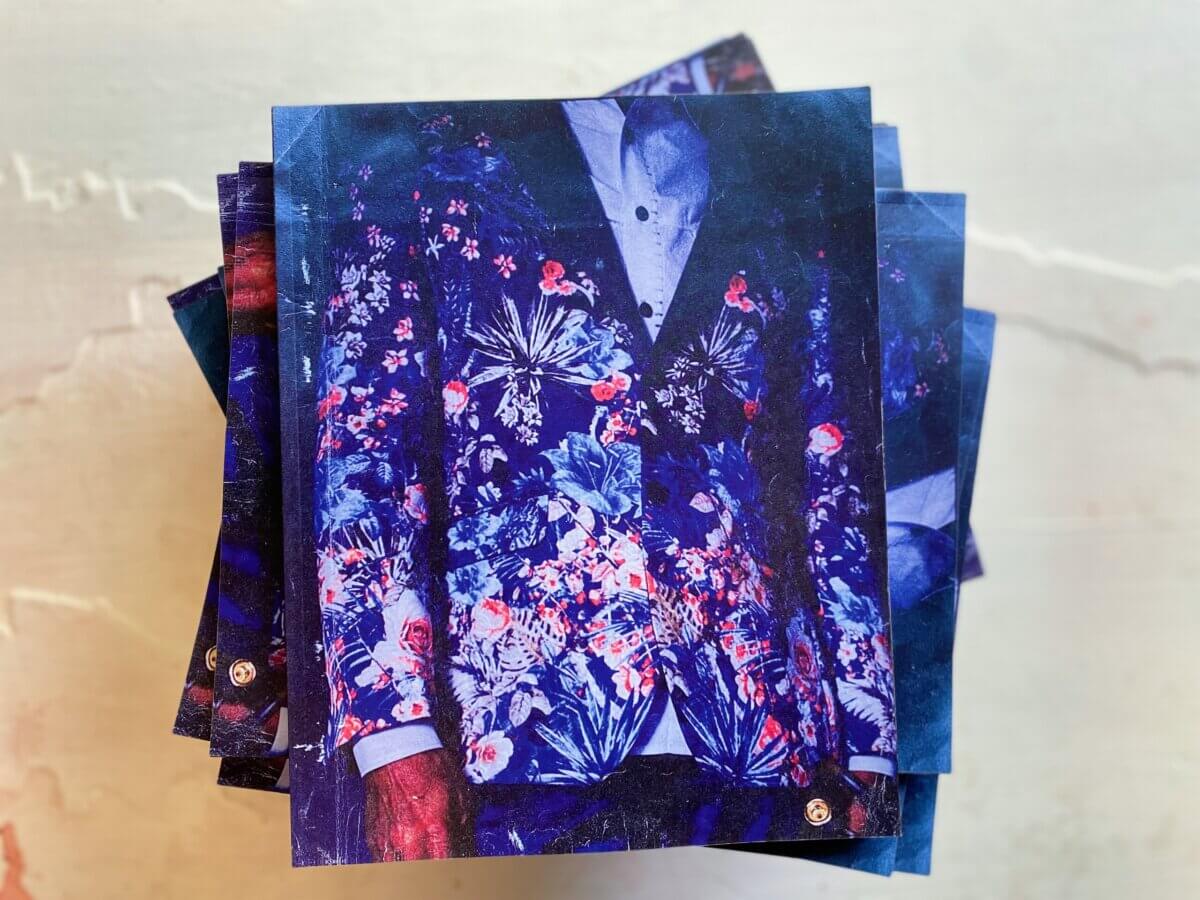 A stack of "An Alabama Song" books with a cover featuring a person wearing a suit jacket with a vibrant blue and red floral pattern over a light shirt, created by Alabama artist Tara Stallworth Lee.