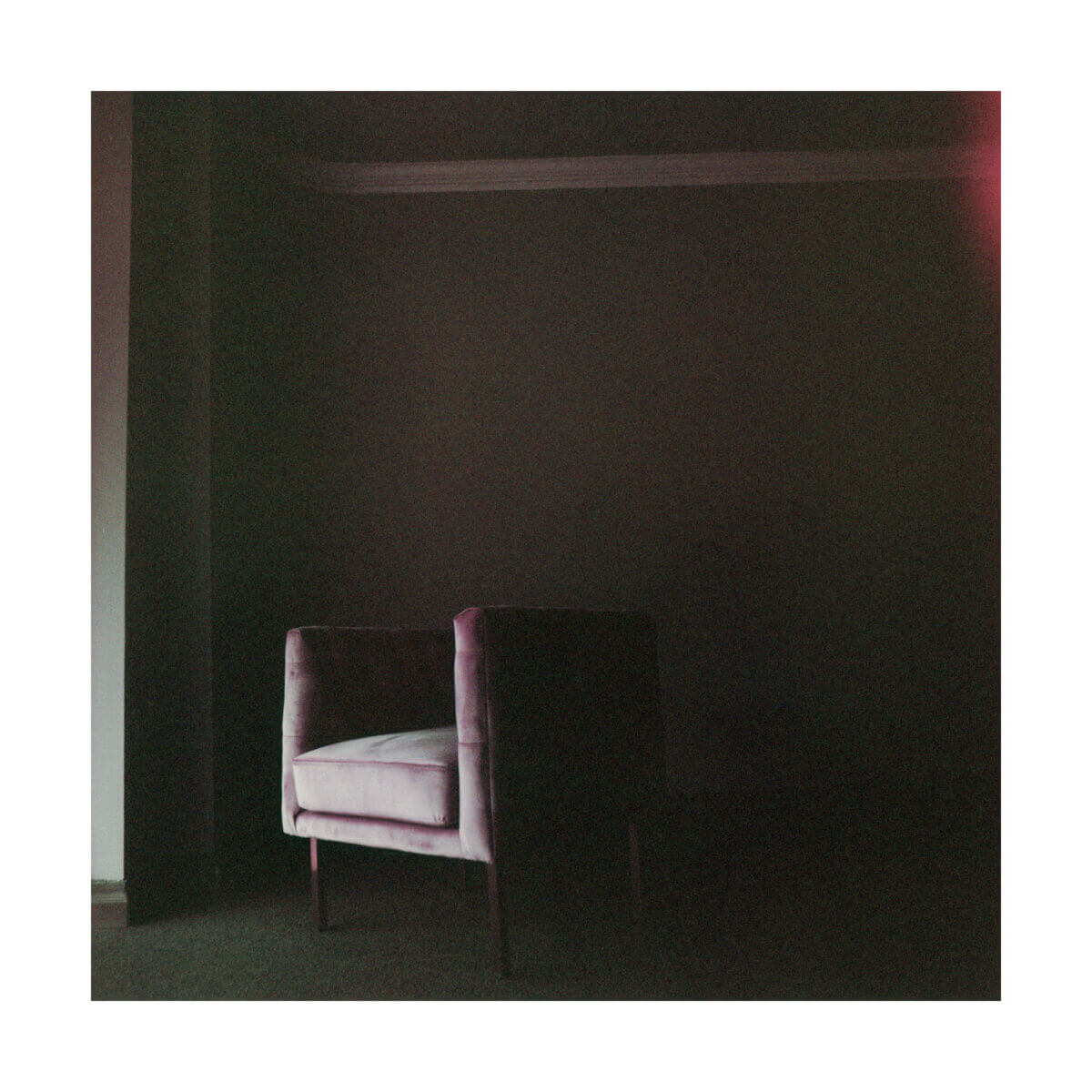 A single upholstered armchair sits in a dimly lit room with dark walls, illuminated by faint, soft light from the left.