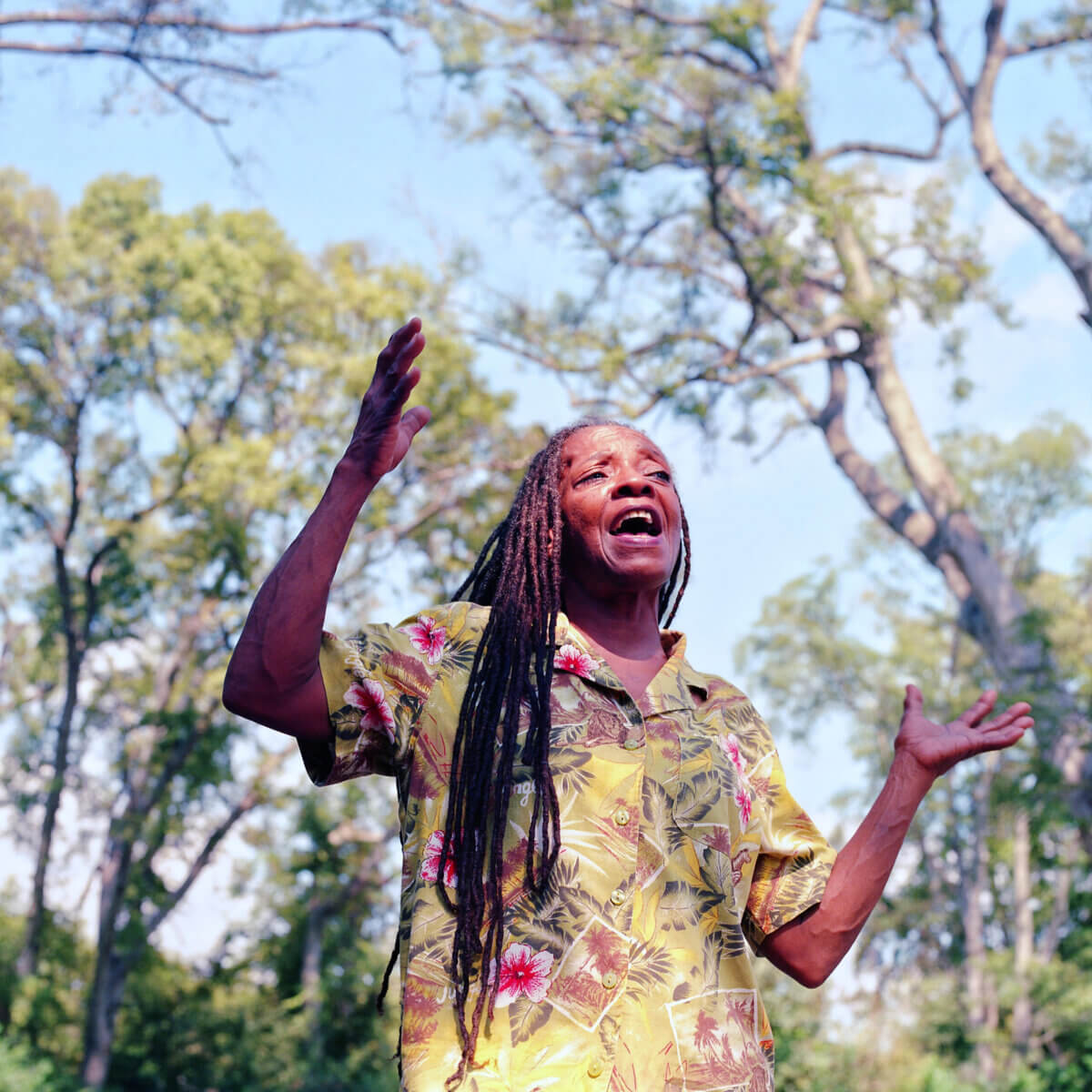 Elaine Hudson with long hair wearing a yellow floral shirt gestures with raised hands outdoors, surrounded by tall trees under a blue sky.