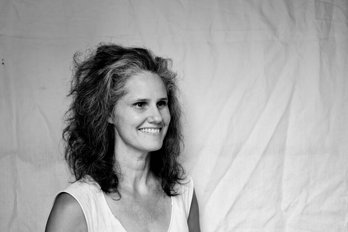 Tara Stallworth Lee with curly hair smiles while standing in front of a plain, light-colored fabric backdrop.