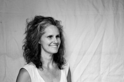 Tara Stallworth Lee with curly hair smiles while standing in front of a plain, light-colored fabric backdrop, embodying the warm spirit of Alabama artist Tara Stallworth Lee.
