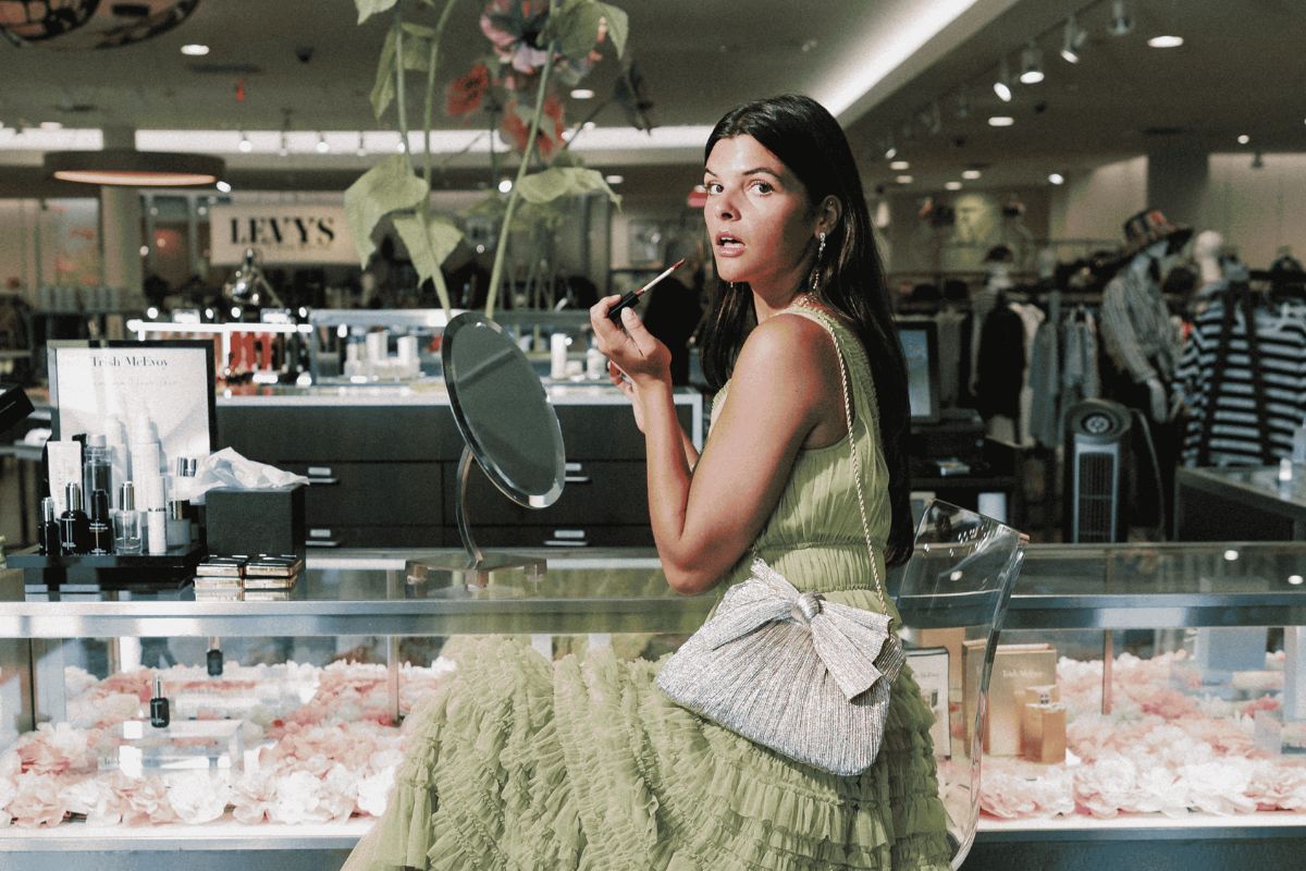 A woman in a green dress sits at a makeup counter in Gus Mayer, Birmingham, applying lipstick while looking in a mirror. She has a textured bag with a large bow.