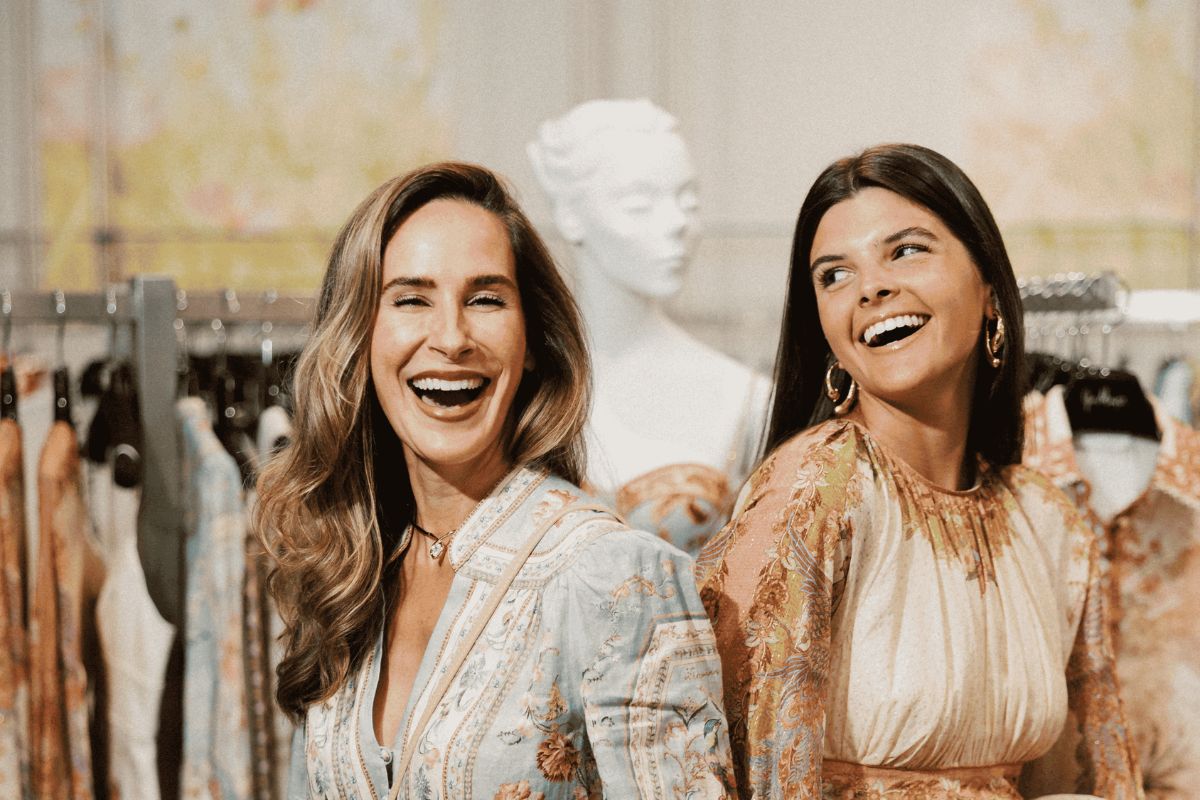 Two women in patterned dresses stand back-to-back, smiling and looking to the side, with racks of clothes and a white mannequin in the background at Gus Mayer in Birmingham.