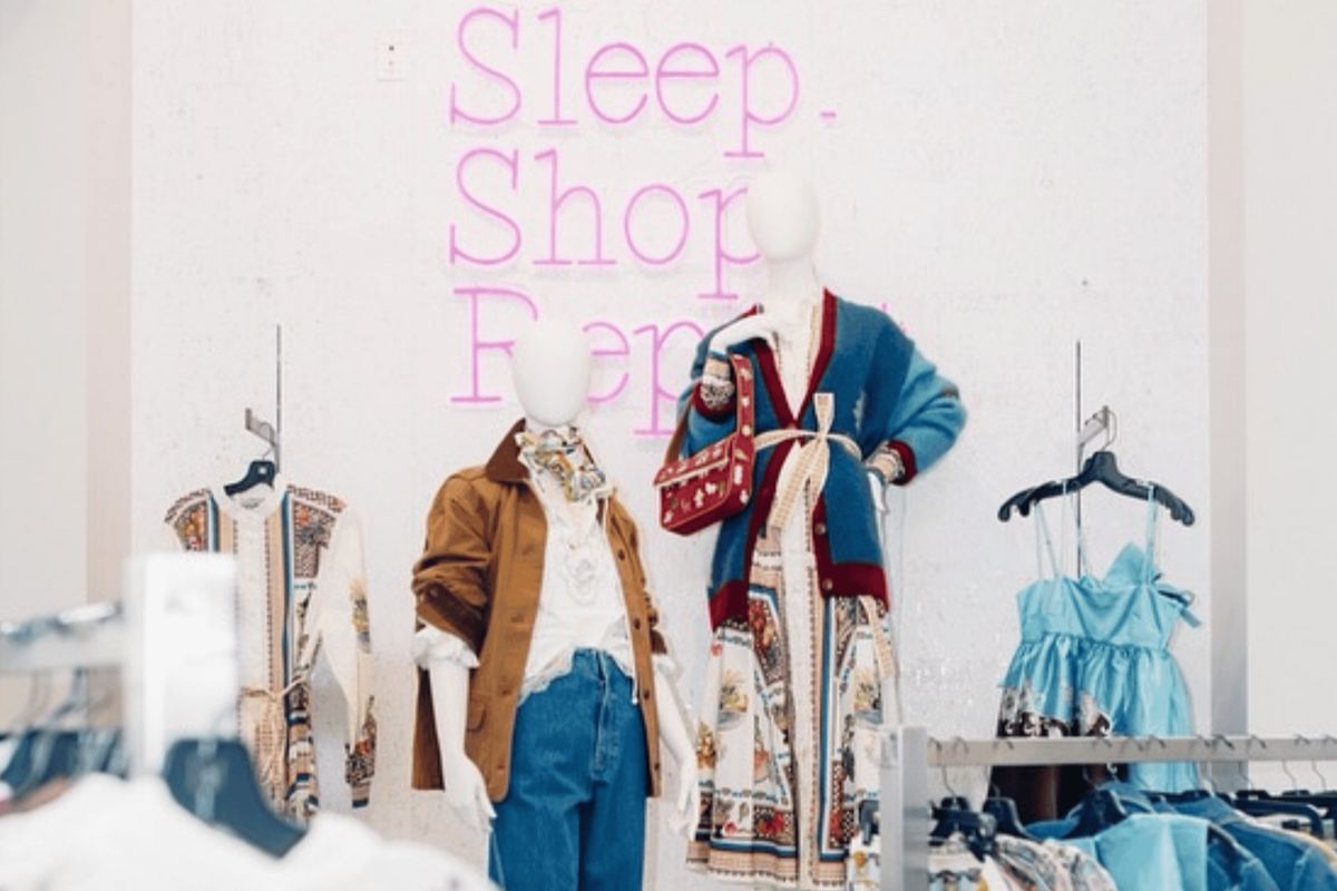 Two mannequins in stylish outfits stand in the Birmingham clothing store Gus Mayer, with a pink neon “Sleep. Shop. Repeat.” sign on the wall behind them and dresses hanging nearby.