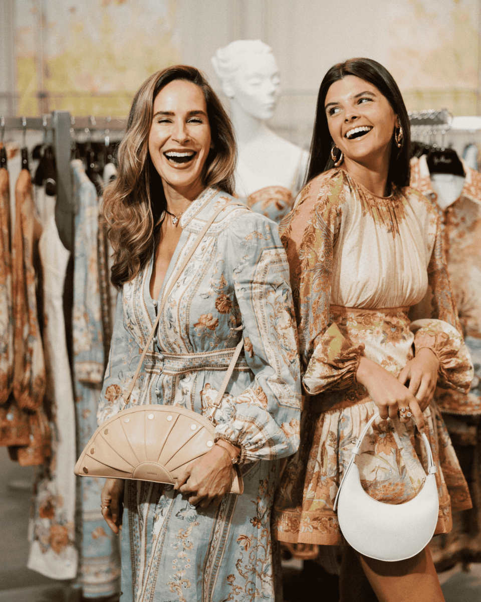 Two women wearing patterned dresses stand and smile, each holding a handbag, in a boutique with clothes racks and a mannequin in the background.