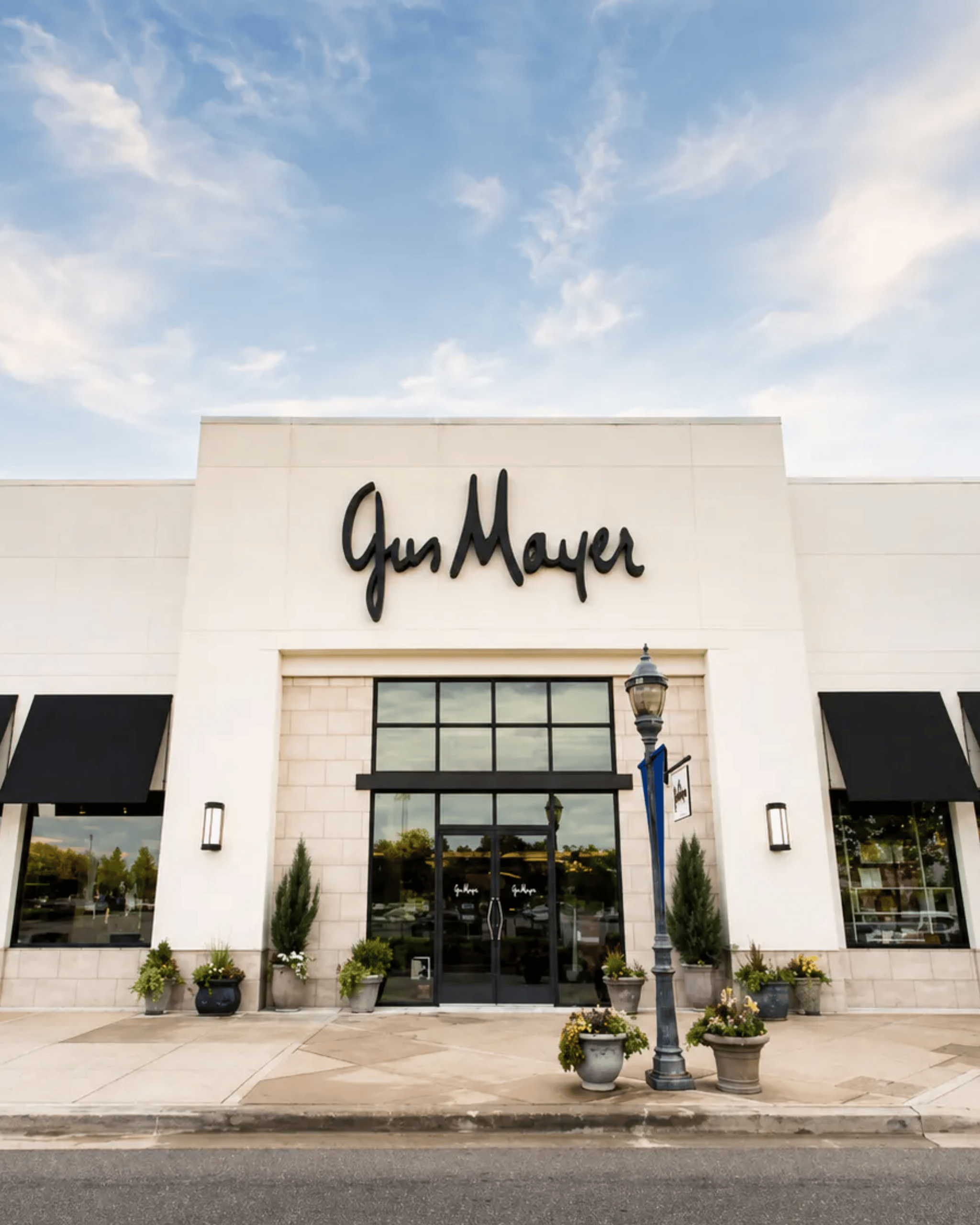 The exterior of a Gus Mayer retail store with black awnings, potted plants by the entrance, and a streetlamp in front under a partly cloudy sky.