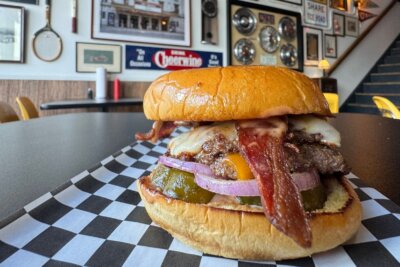 A cheeseburger with bacon, pickles, onions, and melted cheese sits on checkered paper in a diner with framed pictures and car parts on the wall—a classic scene reminiscent of some of the vibrant new restaurants in Birmingham.