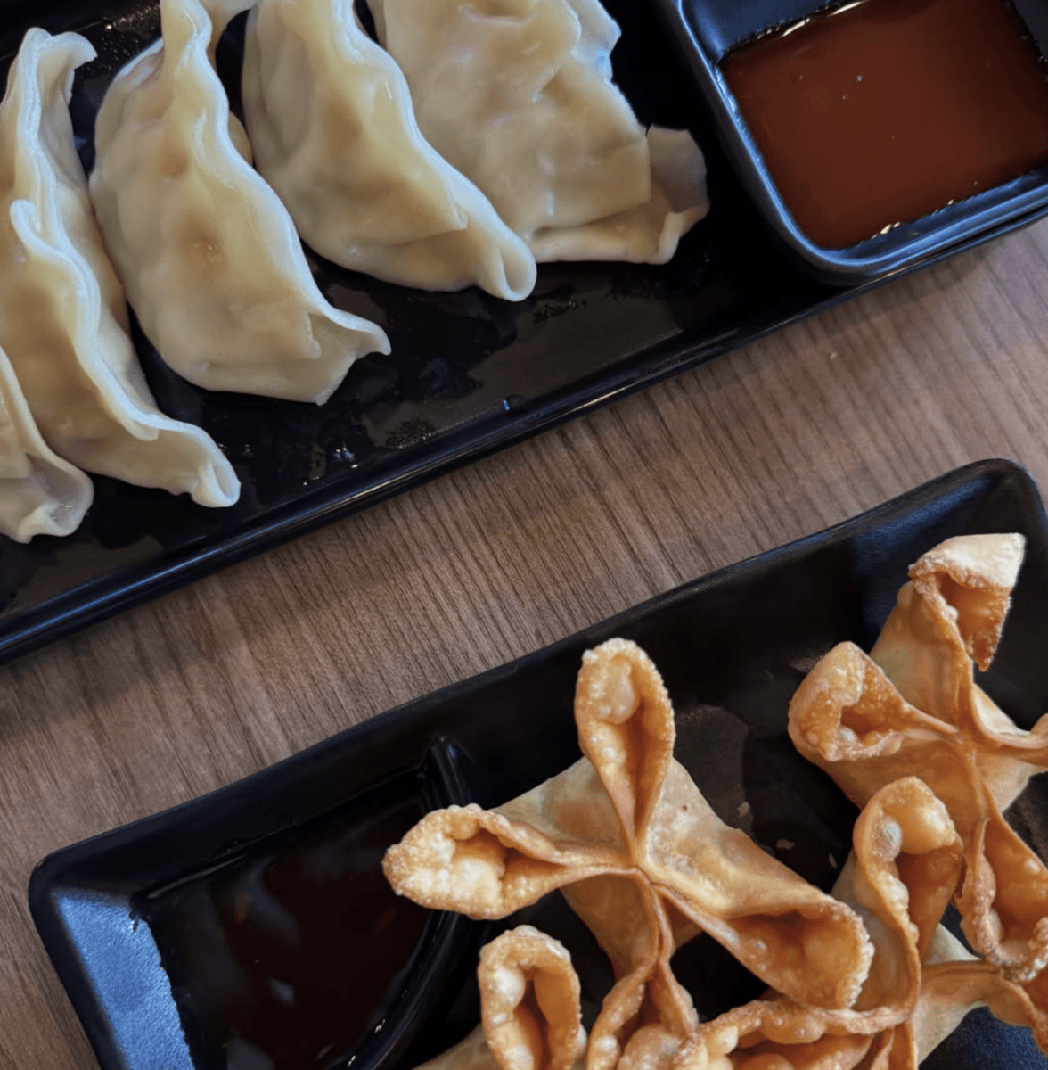 Two black plates with steamed dumplings and fried wontons, each served with a side of dipping sauce, on a wooden table—a delicious example of the creative offerings you might find at new restaurants in Birmingham.