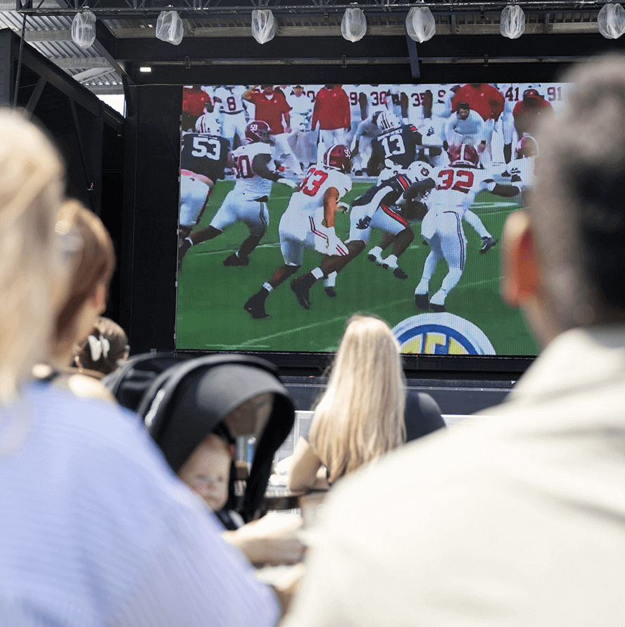 People sitting outdoors watching a football game on a large screen, with players in red and white uniforms visible on the screen.