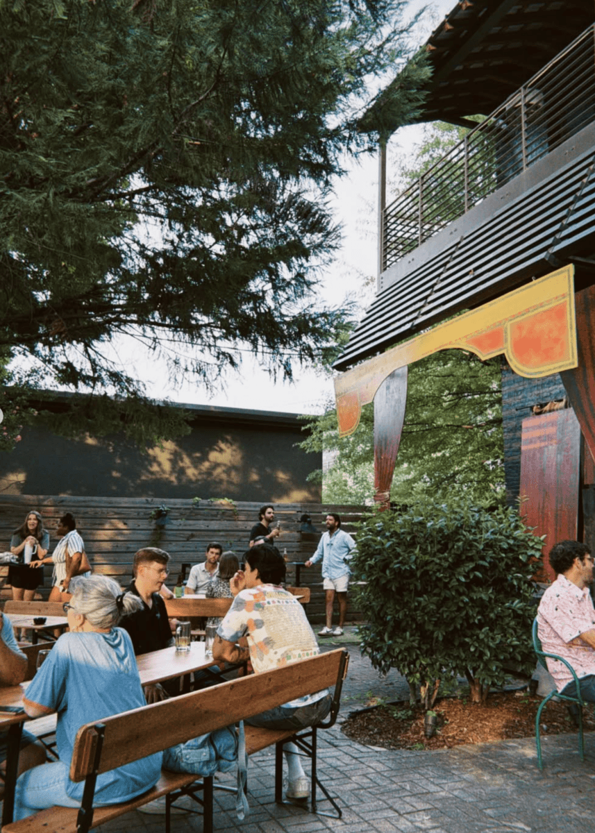 People enjoy outdoor dining as they sit at wooden tables and benches, talking and eating under trees near a building with a balcony on a sunny day.