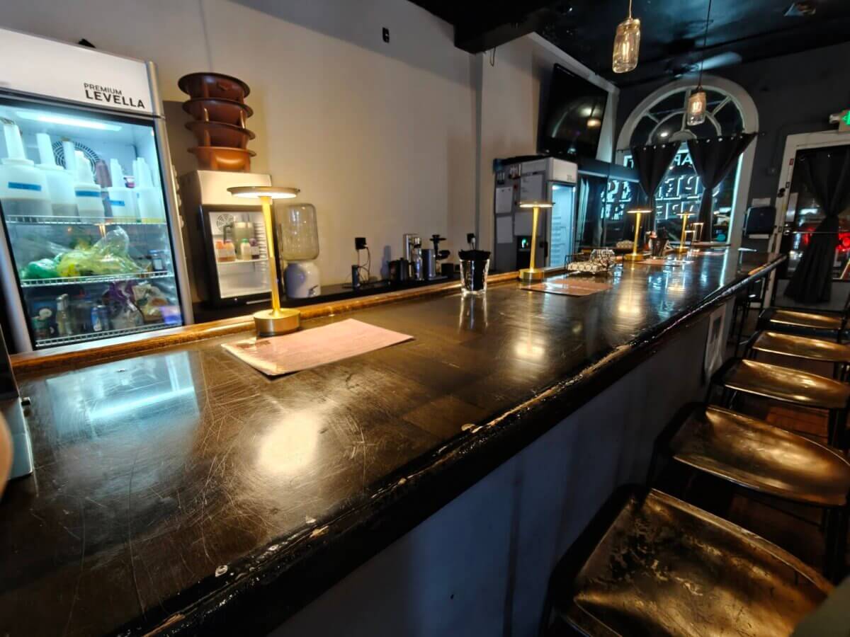 Long wooden bar counter with several stools, countertop lamps, and a refrigerator stocked with drinks and food in the background. The lighting is dim and the space appears empty.