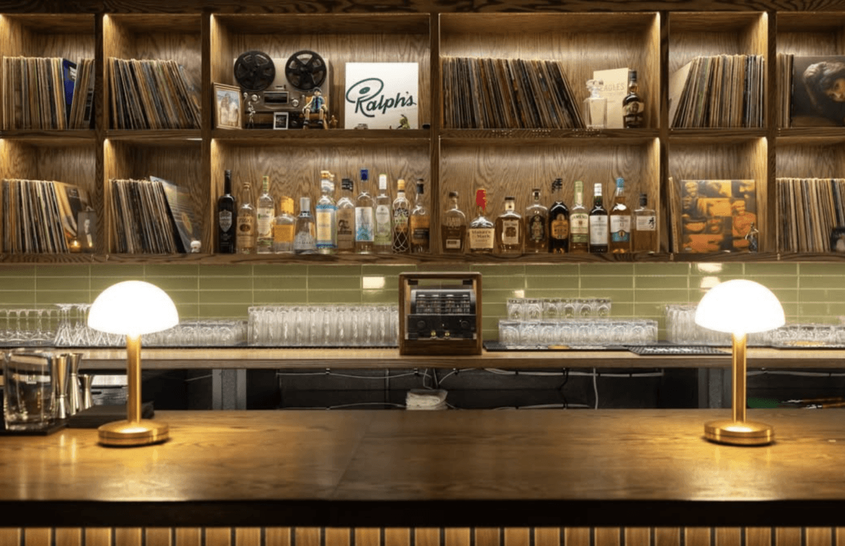 A bar with a wooden counter, two lit lamps, shelves of vinyl records, bottles of liquor, and glassware, with a green tiled backsplash—an inviting scene.