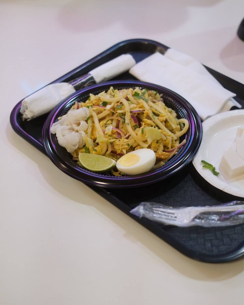 A black tray holds a plate of noodles with vegetables, half a boiled egg, lime wedge, crackers, a wrapped napkin, a fork, and a small plate with tofu cubes.