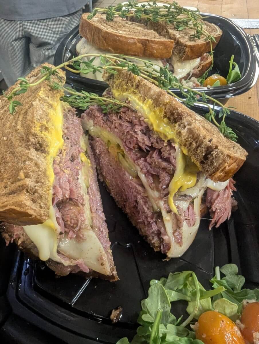 Enjoy classic dining with a sandwich featuring thick slices of rye bread, corned beef, melted cheese, mustard, and herbs, served on a black tray alongside fresh greens and cherry tomatoes—a standout option among new Birmingham restaurants.
