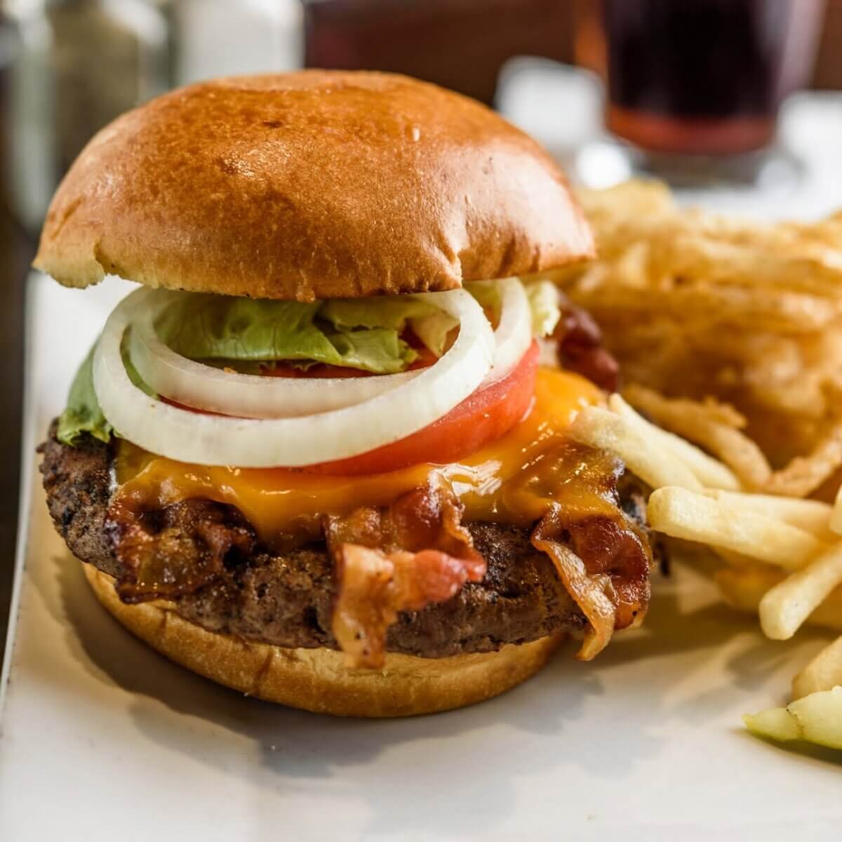 A bacon cheeseburger with lettuce, tomato, and onion on a bun, served with French fries and onion rings on a white plate—perfect for enjoying at BHAM Bars or Breweries while watching football with friends.