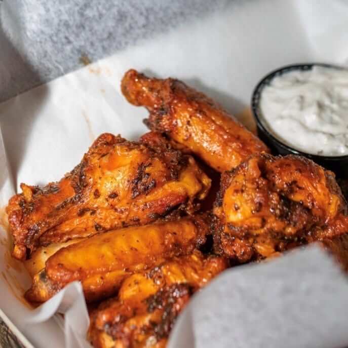 A close-up of seasoned chicken wings in a paper-lined basket, served with a side of creamy dipping sauce—the perfect snack for watching football at your favorite BHAM Bars or local breweries.