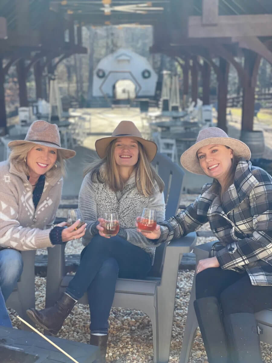 Three women in hats sit outdoors on chairs, smiling and holding drinks at one of Birmingham’s gorgeous wineries, with a wooden pavilion and a large, white geometric structure in the background.