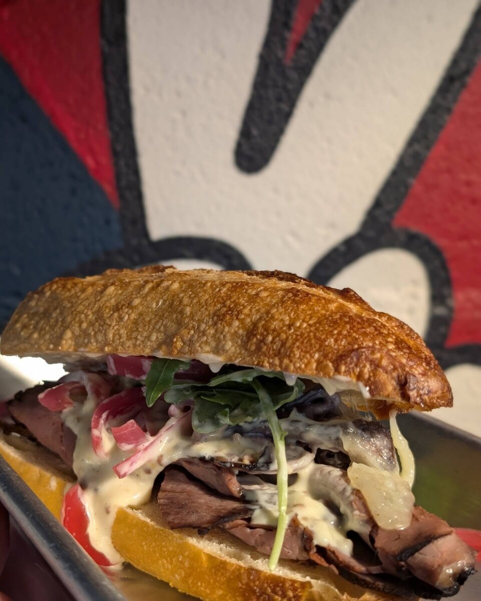 A close-up of a deli sandwich with roast beef, arugula, tomatoes, onions, and sauce on crusty bread, placed on a metal tray with a colorful mural in the background—this is one of BHAM Deli Sandwiches’ epic sandwiches.