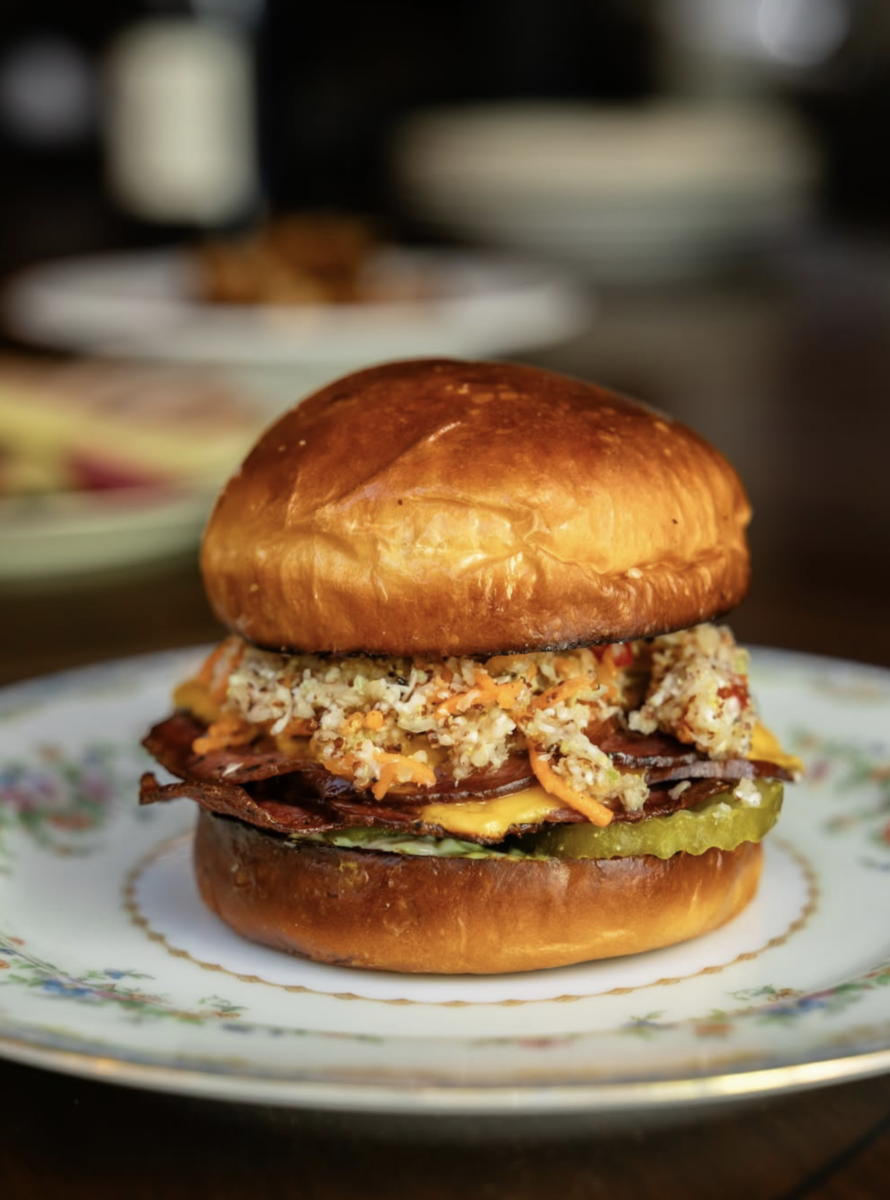 A delicious Epic BHAM Deli Sandwiches creation sits on a floral plate, featuring a toasted bun piled high with pickles, bacon, shredded coleslaw, and tangy sauce.