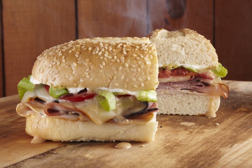 A sesame seed sandwich roll filled with sliced deli meat, melted cheese, lettuce, tomato, and mayonnaise—cut in half on a wooden surface—this is one of our Epic Sandwiches from the BHAM Deli Sandwiches collection.