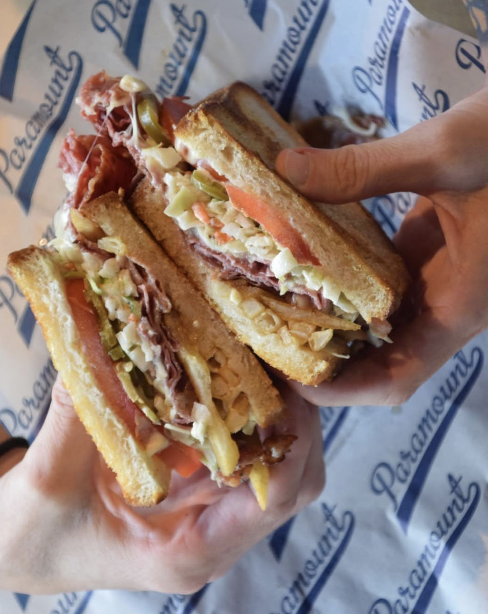 Two hands hold thick, epic sandwiches—stacked high with sliced bread, deli meat, tomato, lettuce, and melted cheese. The delicious deli sandwiches rest on Paramount-branded paper.