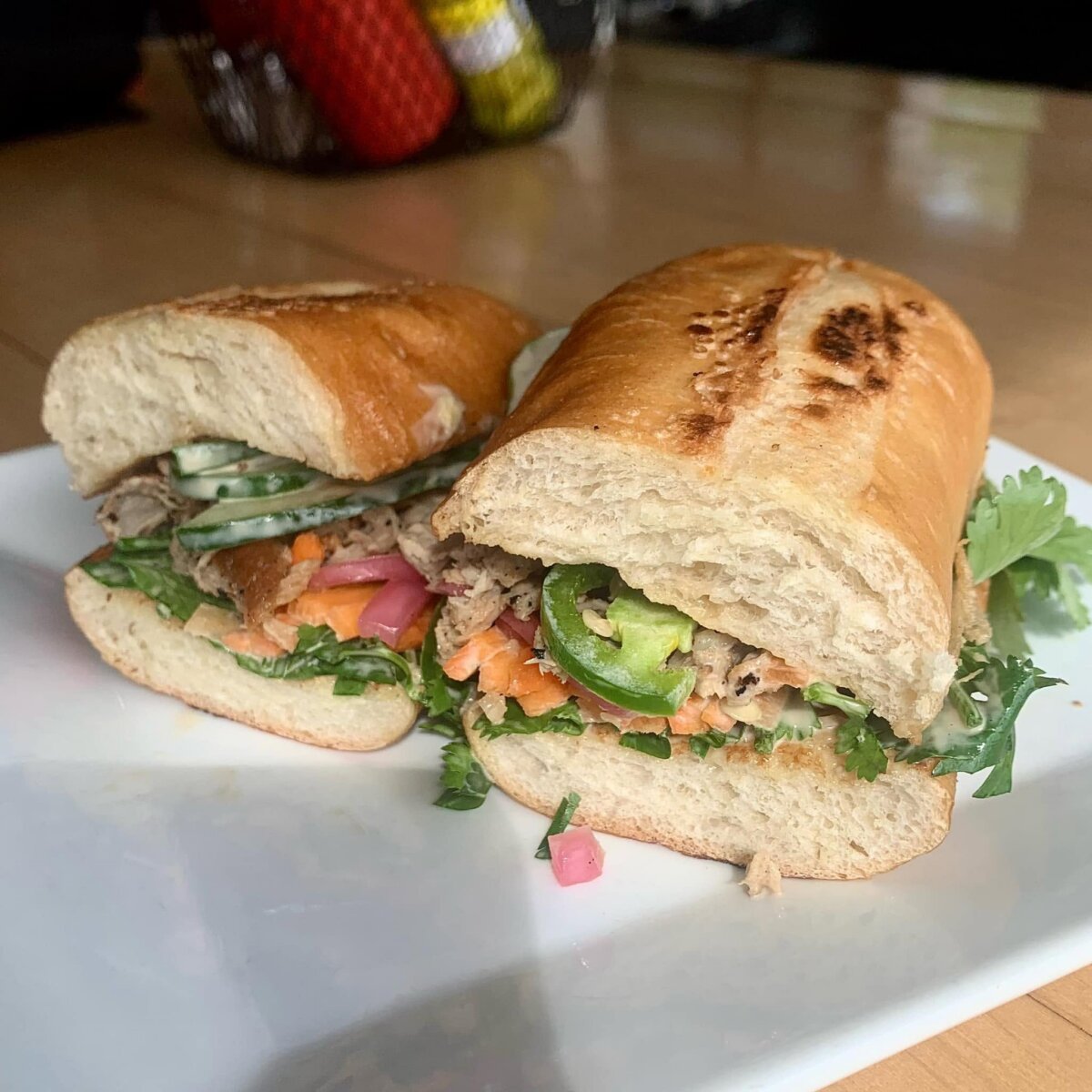 A banh mi sandwich cut in half on a white plate, filled with vegetables, herbs, and meat, stands out among Epic Sandwiches, with a basket of condiments in the background.