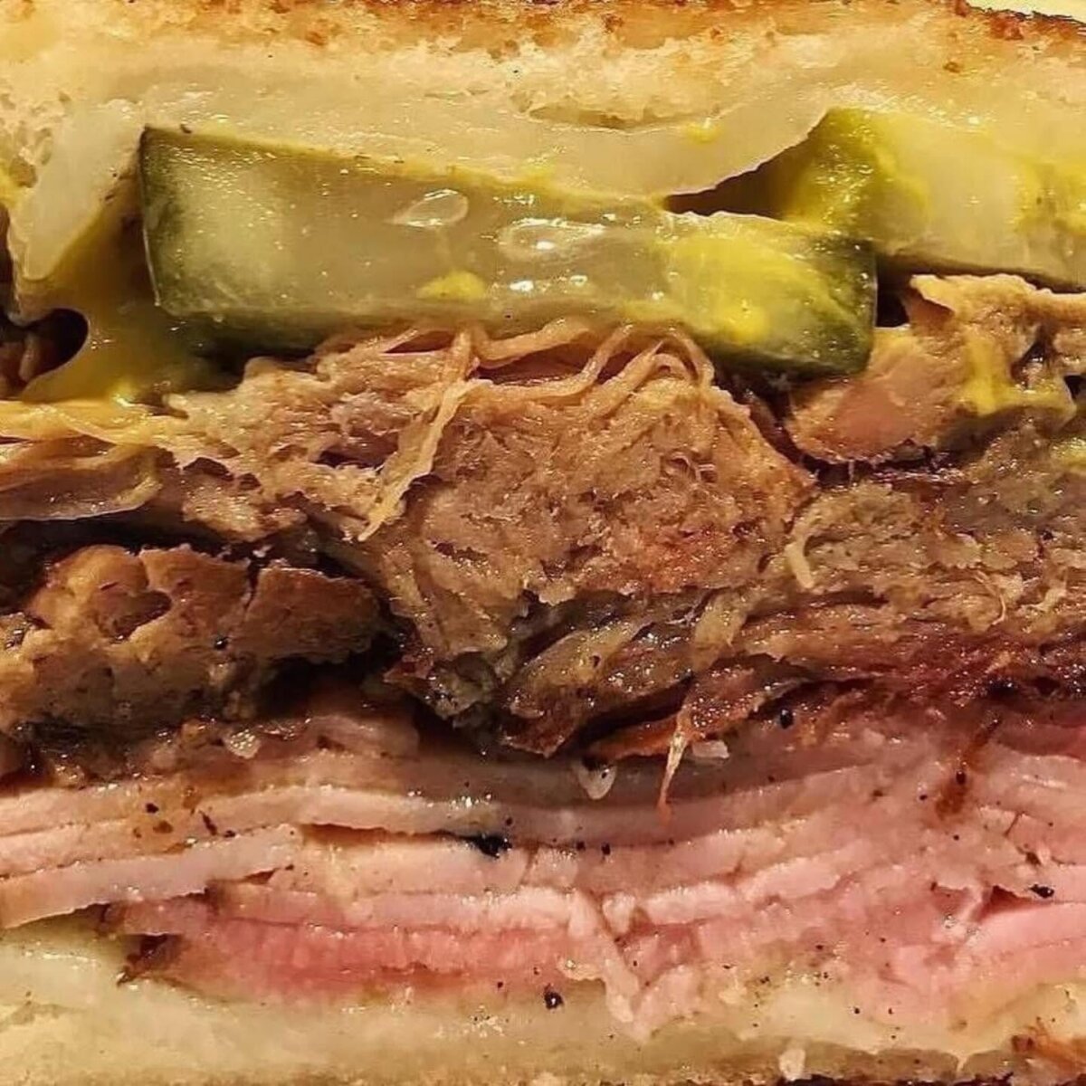 Close-up of a sandwich showing layers of sliced ham, shredded pork, pickles, melted cheese, mustard, and toasted bread—an epic sandwich inspired by the deli sandwiches at BHAM Deli.